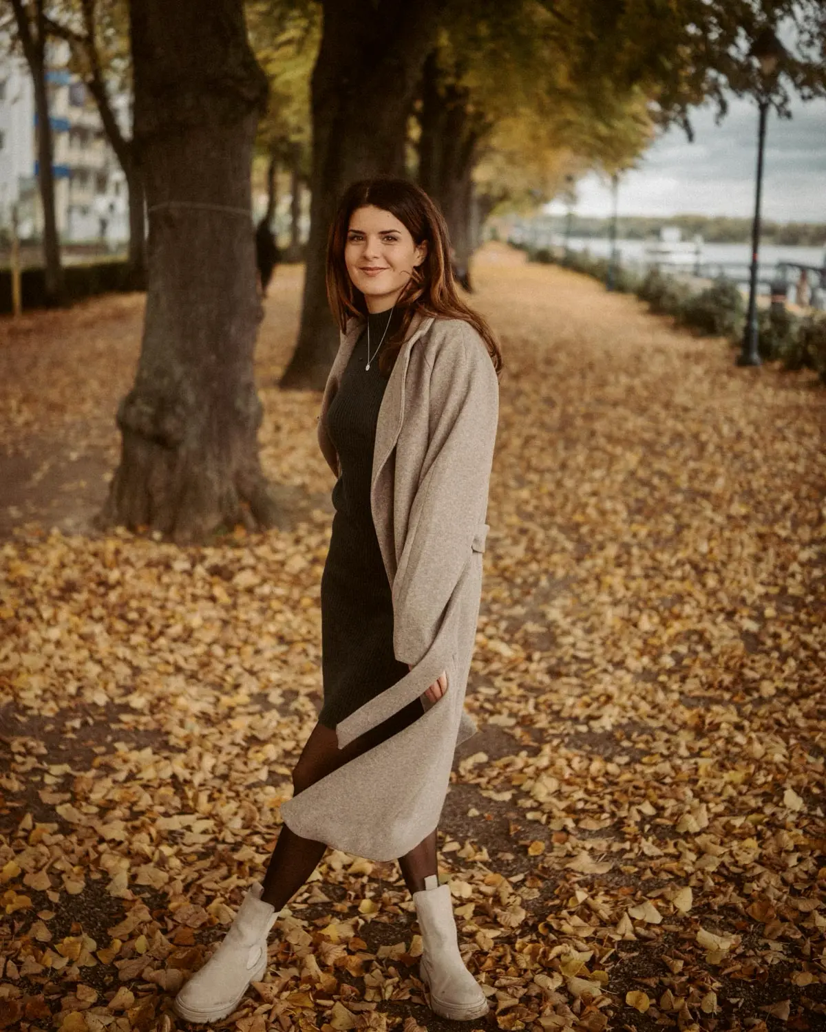 Frau steht auf einem Weg voller Herbstlaub, Mantel offen und Blick ruhig nach vorn