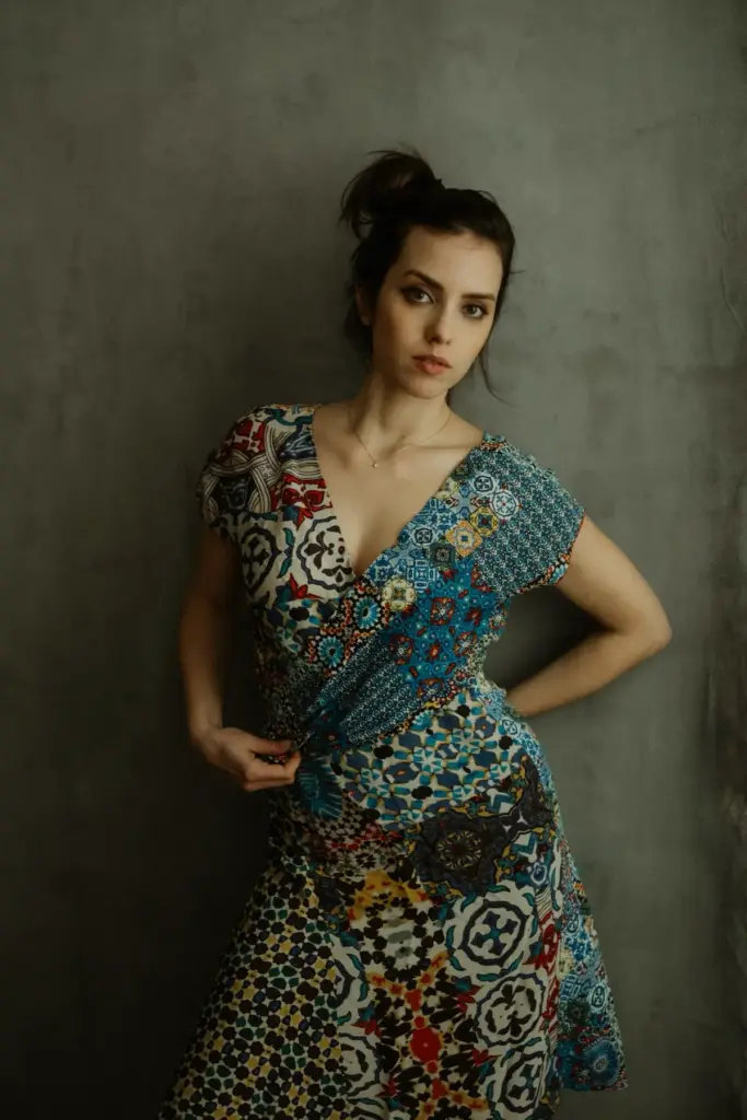 Frau im gemusterten Kleid lehnt an einer grauen Wand und schaut offen in die Kamera