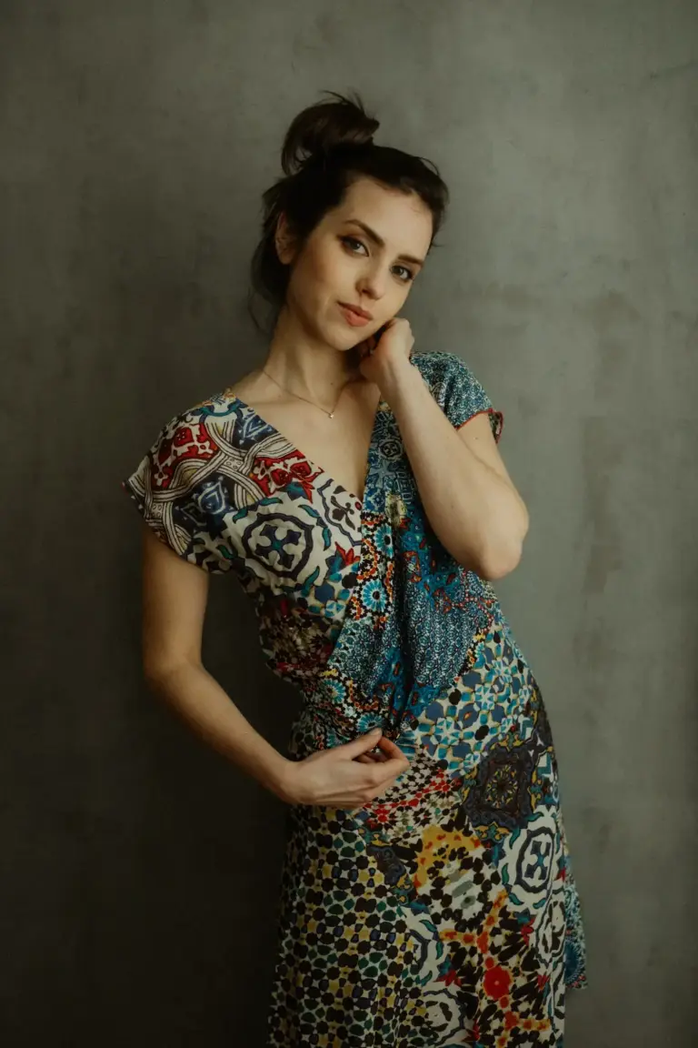 Frau im gemusterten Kleid lehnt an grauer Wand, Blick ruhig und leicht seitlich
