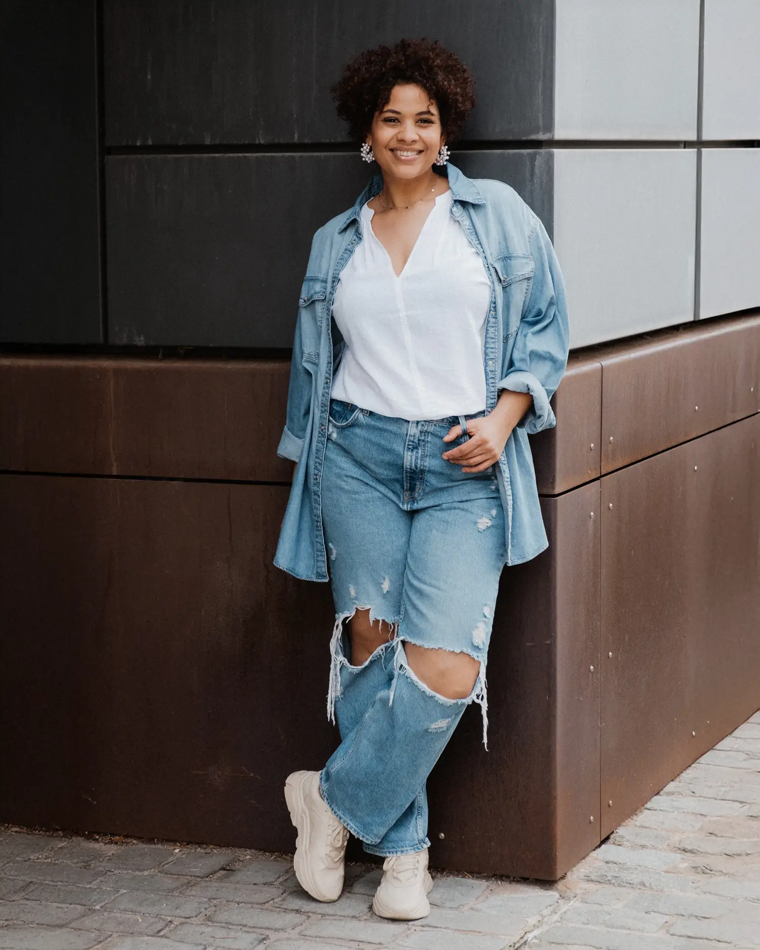 Frau lehnt lächelnd an einer dunklen Gebäudeecke, Jeansjacke locker über dem Outfit