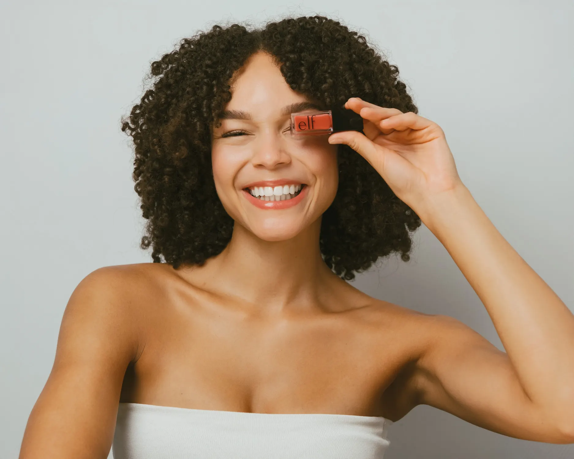 Lächelndes Model mit Locken hält ein Kosmetikprodukt vor ein Auge