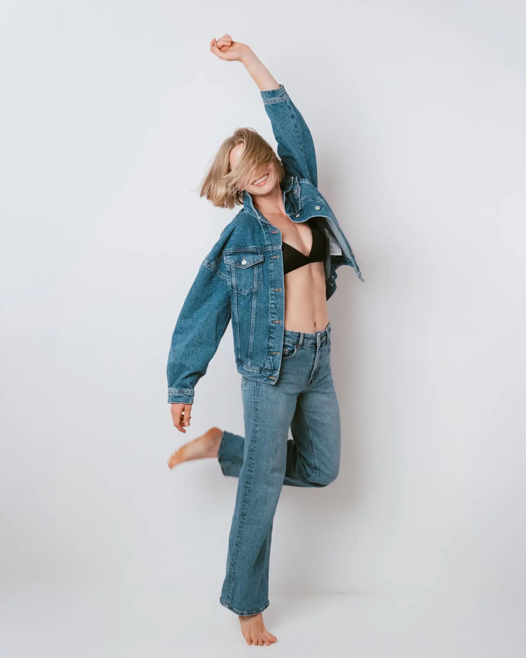 Model in Jeansjacke und Jeans, barfuß mit erhobenem Arm und angewinkeltem Bein vor hellem Hintergrund