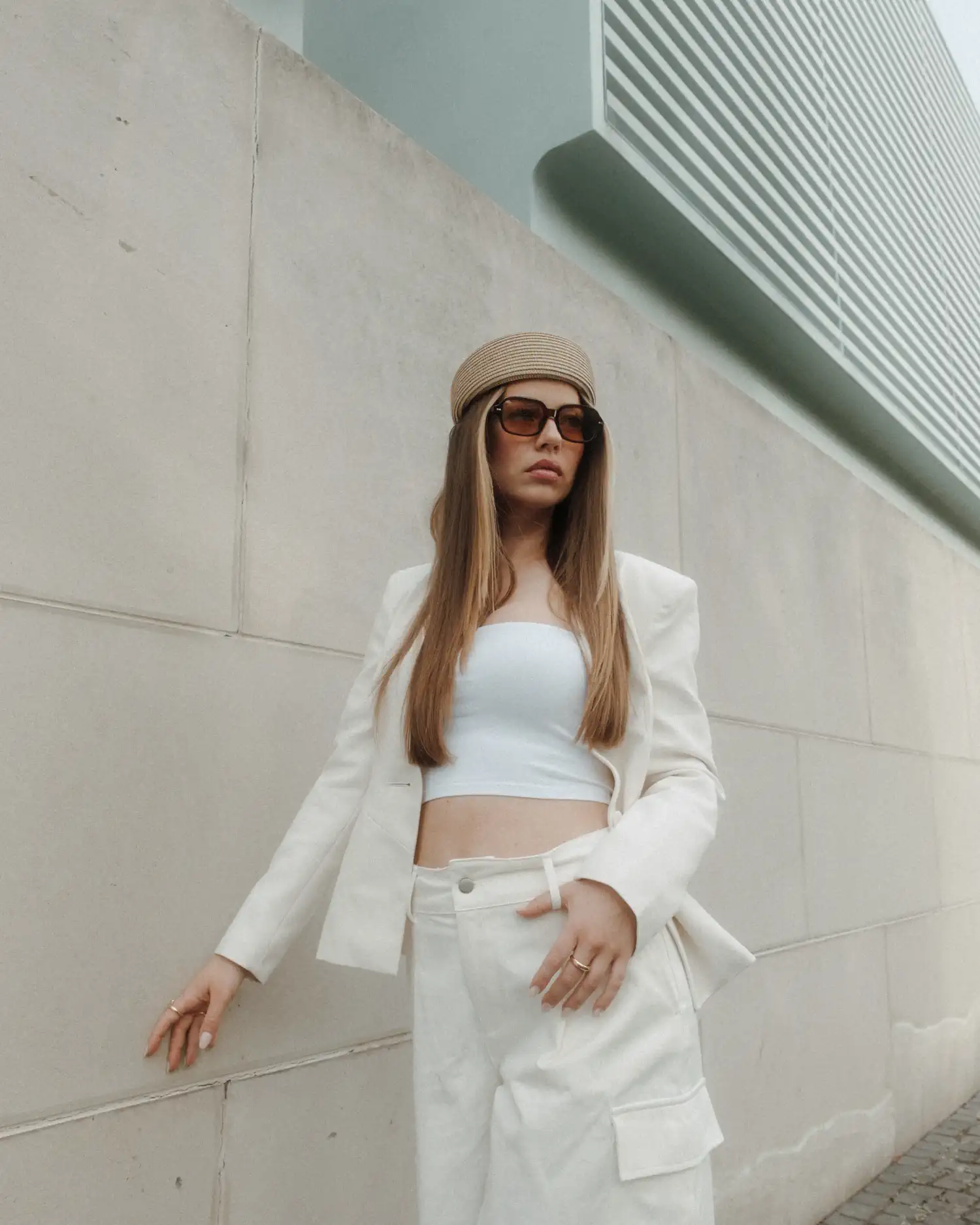 Model in weißem Outfit mit Sonnenbrille und Mütze vor einer Betonfassade