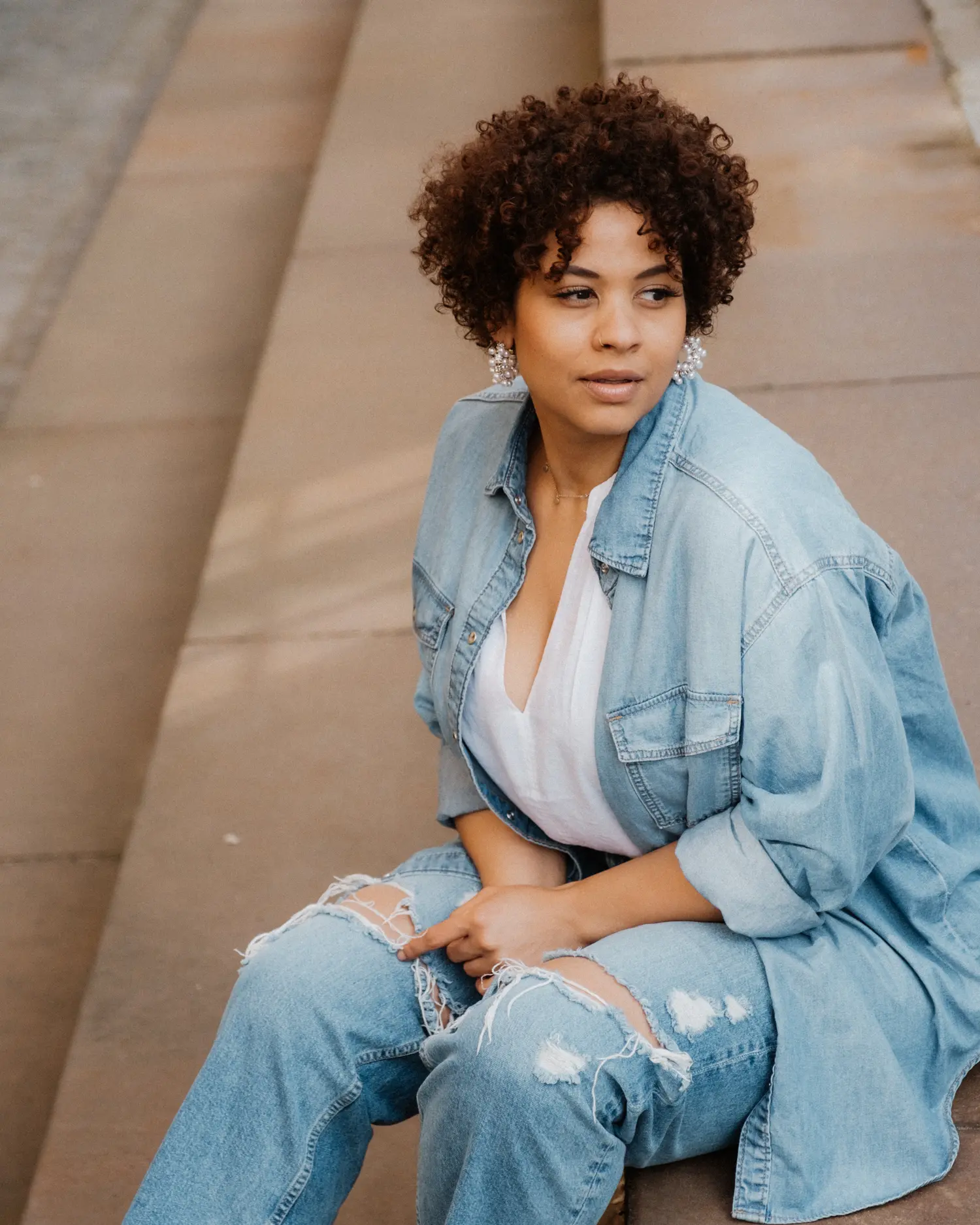 Model mit kurzen Locken in Jeanskleidung, auf einer Steintreppe sitzend