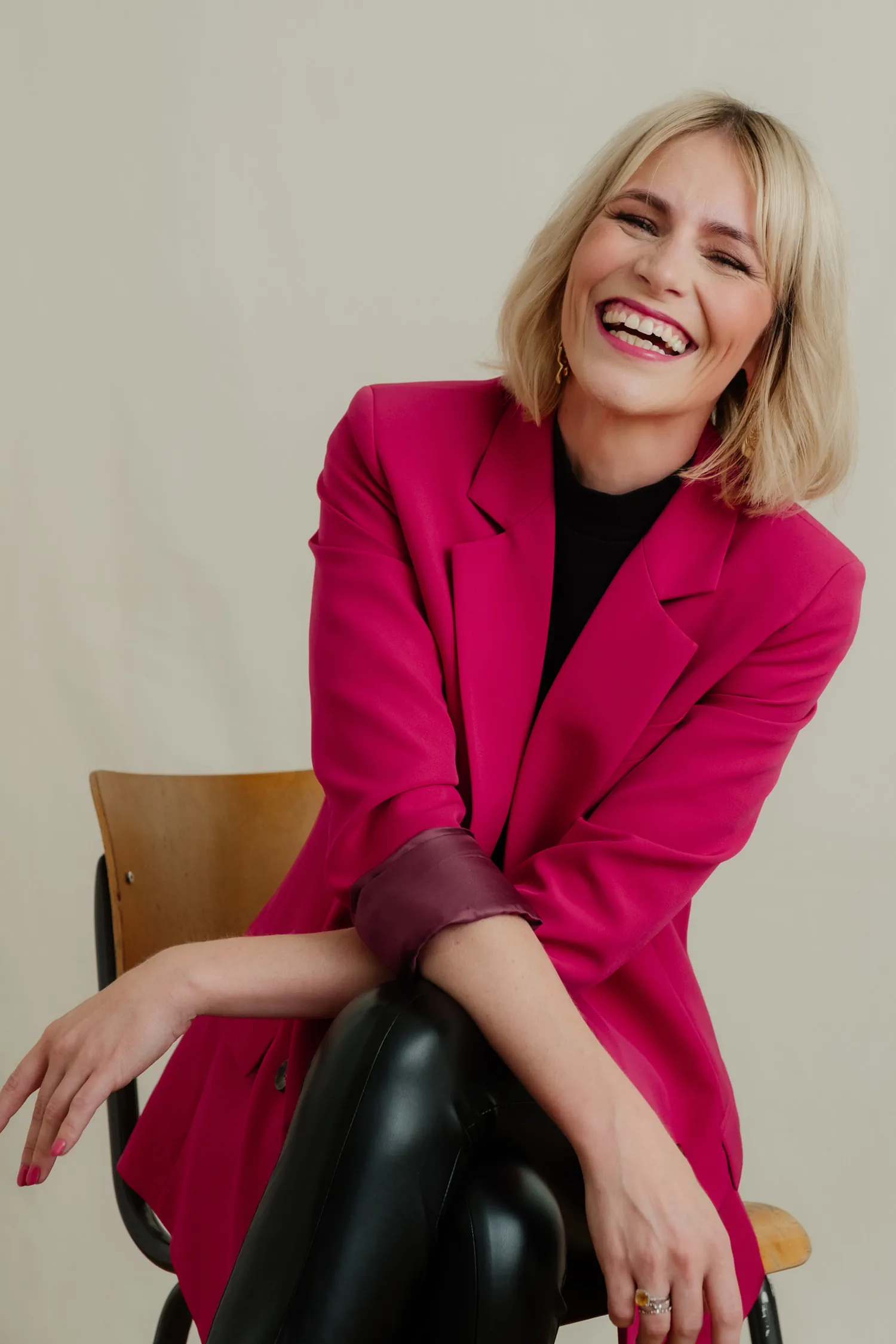 Julia Kleine lachend im pinkfarbenen Blazer auf einem Holzstuhl, schwarze Lederhose