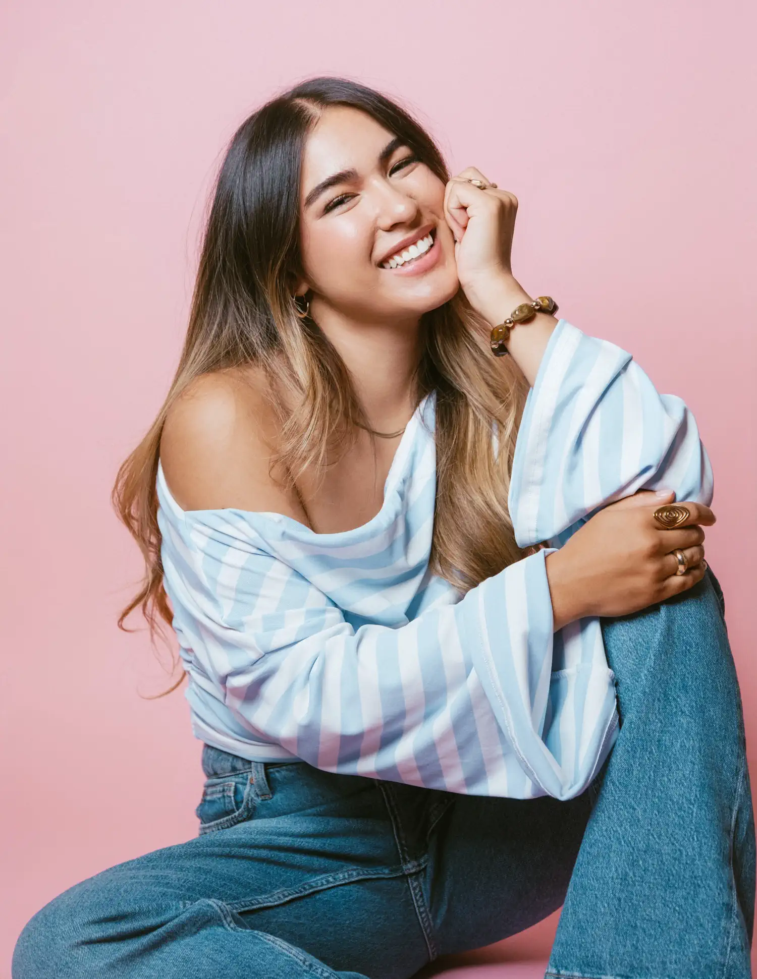Lachendes Model im blau-weiß gestreiften Oberteil, mit einer Hand an der Wange vor rosa Hintergrund