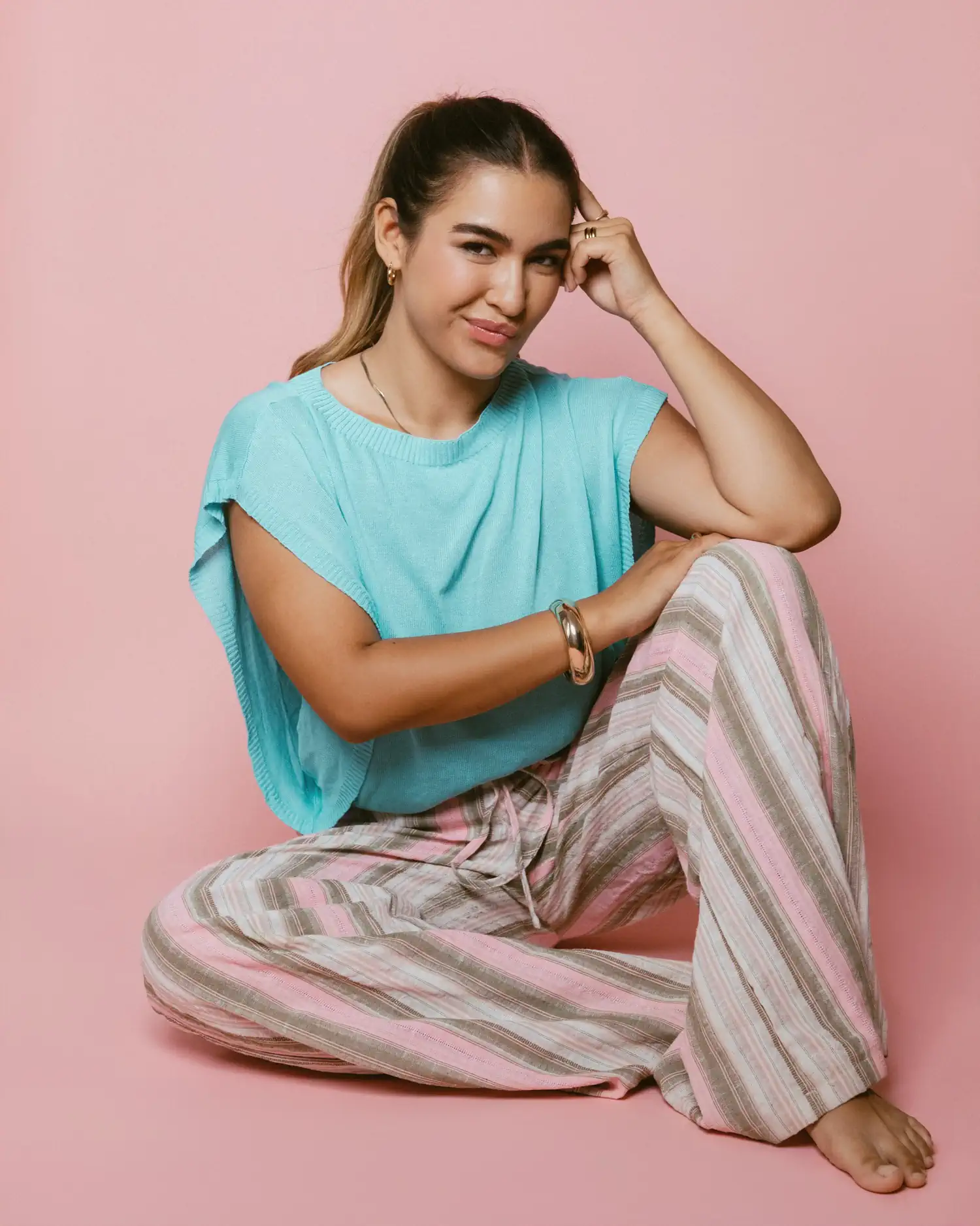 Model im türkisfarbenen Oberteil und gestreifter Hose, auf dem Boden vor rosa Hintergrund sitzend